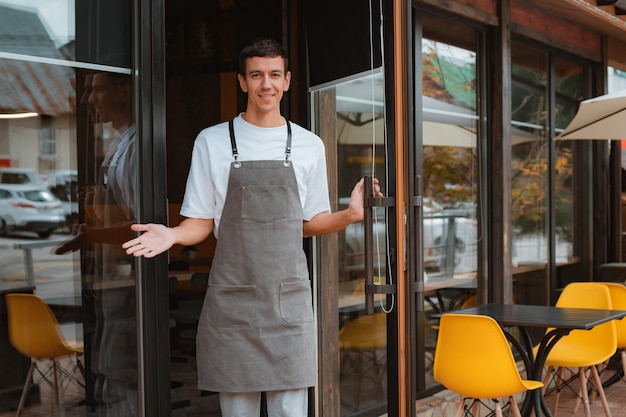 Barista of ober café of coffeeshop eigenaar tegen ingangsgebaar die u uitnodigt om een glimlachende man in schort te bezoeken die buiten staat en trots is op zijn kleine lokale bedrijf