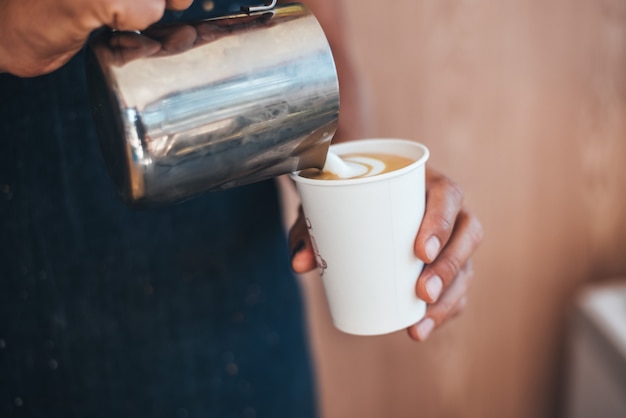 The barista pours milk from a tin can into coffee in a paper disposable cup