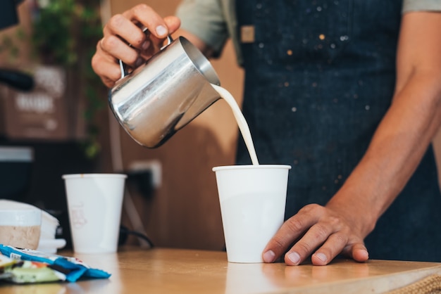 The barista pours milk from a tin can into coffee in a paper disposable cup