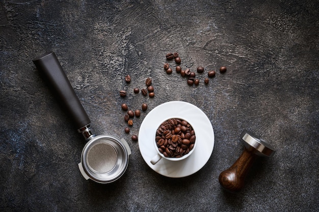 暗い織り目加工の石の背景に豆とバリスタツールとコーヒーカップ、コピースペースのある上面図