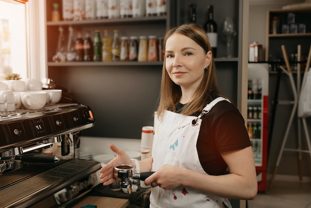 A barista with white apron smiles and poses holding a metal tamper and a portafilter with coffee