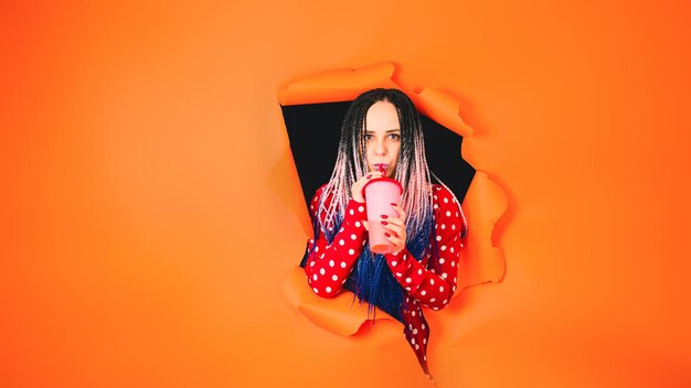 Barmaid with drink looking through hole in paper background Female bartender with dreadlocks and cup of sweet beverage peeking through ripped orange paper background in studio and looking at camera