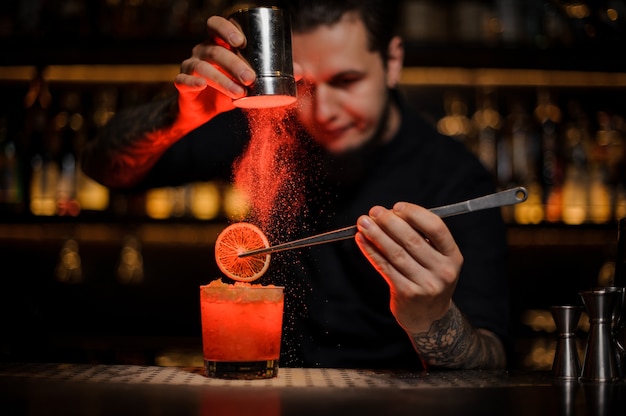 Barman adding to an alcoholic cocktail in the glass a dried orange with tweezers and aromatic powder in the red light on the bar counter