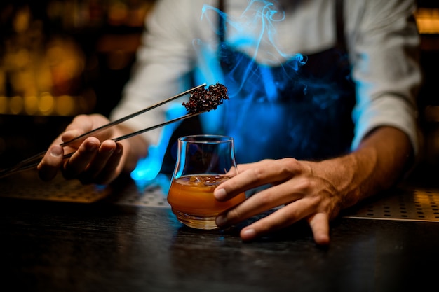 barman adding chilled melting caramel with twezzers to the cocktail glass with ice cubes under blue lights