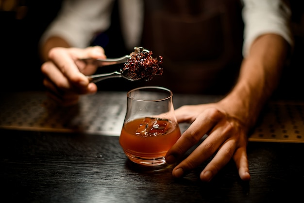 Barman die gekoelde smeltende karamel met twezzers toevoegt aan het cocktailglas met ijsblokjes