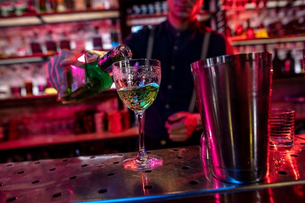 Barman finishes preparation of alcoholic cocktail in multicolored neon light, focus on glass