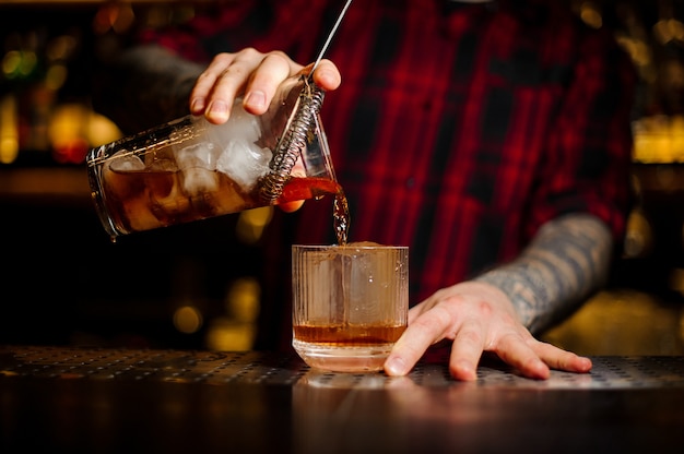 Barman pouring strong alcoholic cocktail of whiskey using strainer into the glass on the bar counter