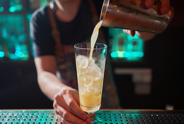 Бармен в рубашке и фартуке делает алкогольный напиток с льдом в коктейльном стакане