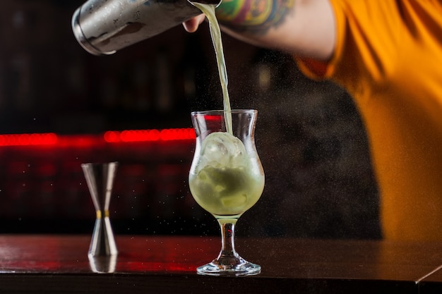 Barman sprenkelt op verlicht glas met felgroene koude cocktail op toog en maakt vuurvlam erover.