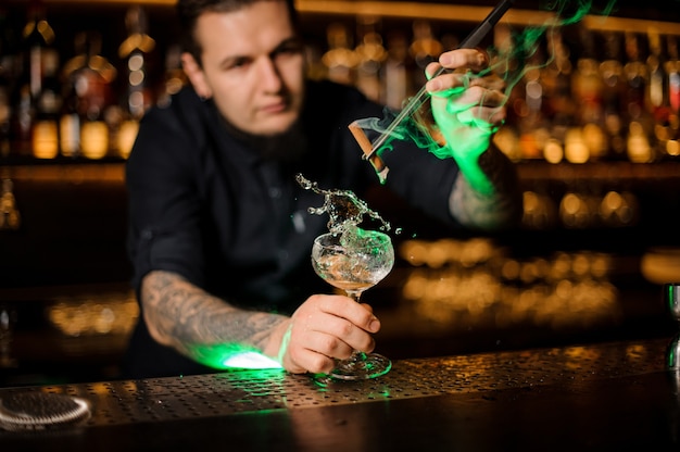 Barman voegt toe aan het cocktailglas met plons met een gedroogde sinaasappel aromatische gerookte kaneel met een pincet op de toog