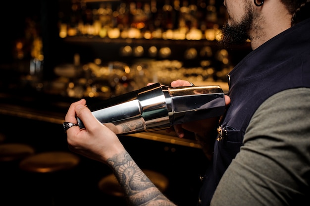 Barman with beard and tattoo on his hand making summer cocktail in shaker