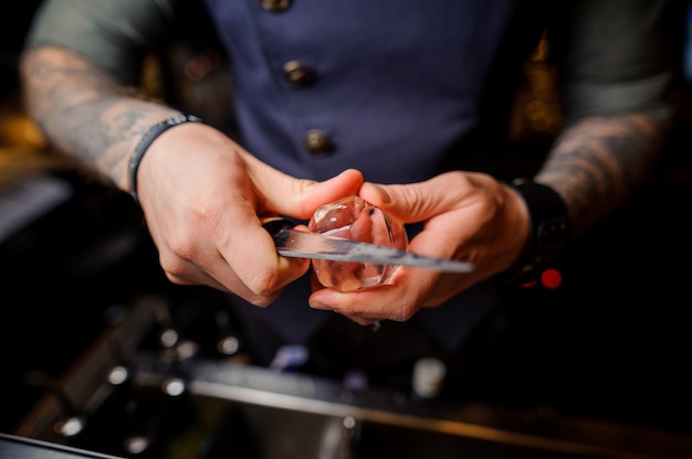 Barman with tattoos on his hands cuts the ice with a special knife