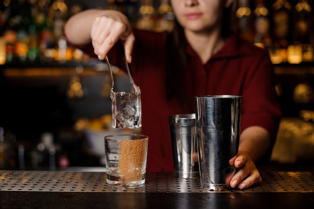 Ragazza del barista che mette un grande cubetto di ghiaccio in un bicchiere vuoto su un bancone bar