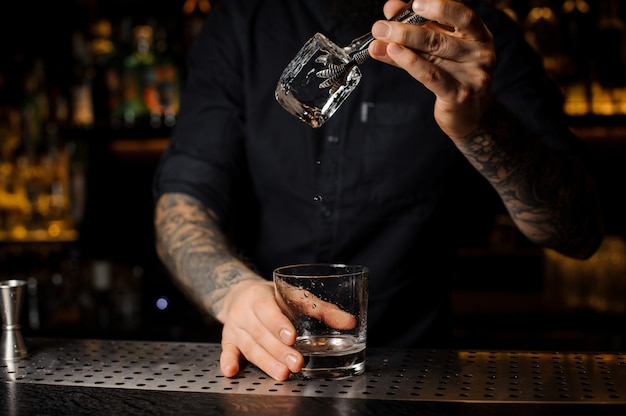 Barista che aggiunge a un drink nel bicchiere un grosso cubetto di ghiaccio con una pinzetta sul bancone del bar