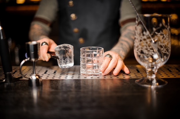 Photo bartender holding a big ice cube near the empty glass