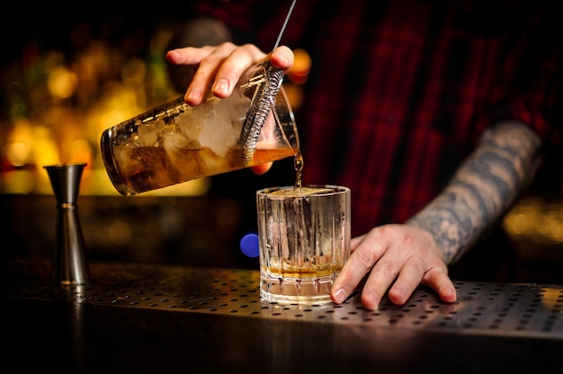 Barista che versa un delizioso cocktail rusty nail dal misurino attraverso il colino a un bicchiere sul bancone del bar