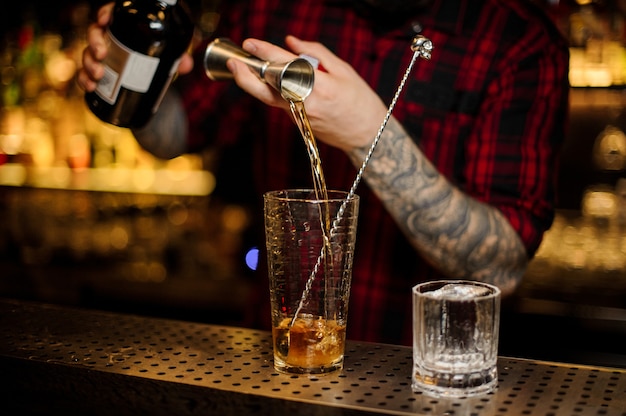 Bartender pouring drink making a Rusty Nail cocktail from the steel jigger to the glass measuring cup on the bar counter