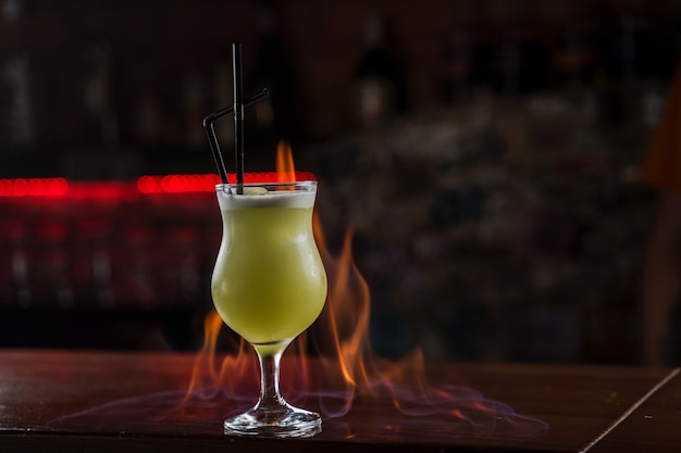 バーテンダーは、バーのカウンターに明るい緑の冷たいカクテルを照らすガラスに振りかけ、その上に火の炎を作ります。