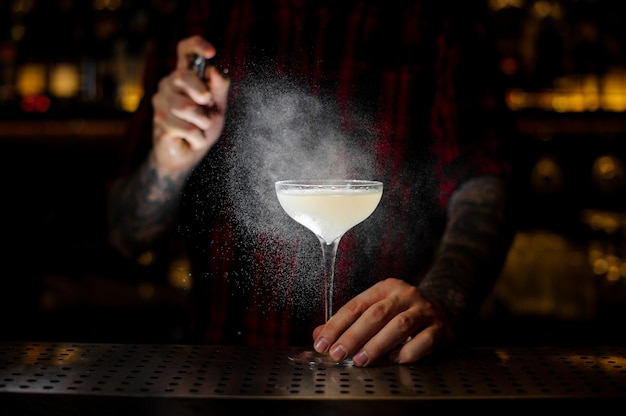 Bartender sprinkling elegant cocktail glass with light and tasty alcoholic drink with bitter on the bar in dark