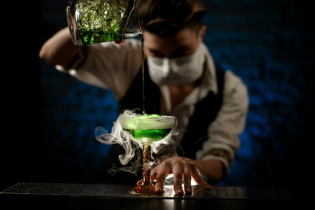 Bartender with medical mask pours cocktail into glass and attentively look at it
