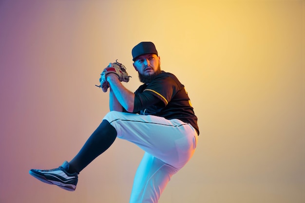 Baseball player, pitcher in a black uniform practicing and training on gradient wall in neon light. Young professional sportsman in action and motion. Healthy lifestyle, sport, movement concept.