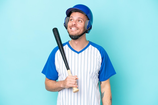 파란색 배경에 격리된 헬멧과 방망이를 든 야구 선수는 올려다보는 동안 아이디어를 생각합니다.