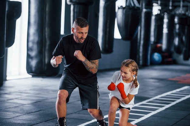 Basic punches. Young tattooed boxing coach teaches cute little girl in the gym.