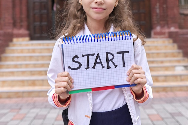 Basisschool meisje student met Kladblok en handgeschreven woord start, schoolgebouw achtergrond. Terug naar school, onderwijsconcept