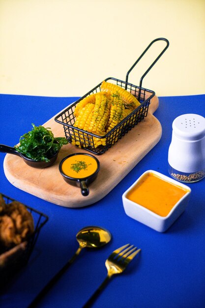 A basket of corn on the cob sits on a table next to a bowl of sauce.