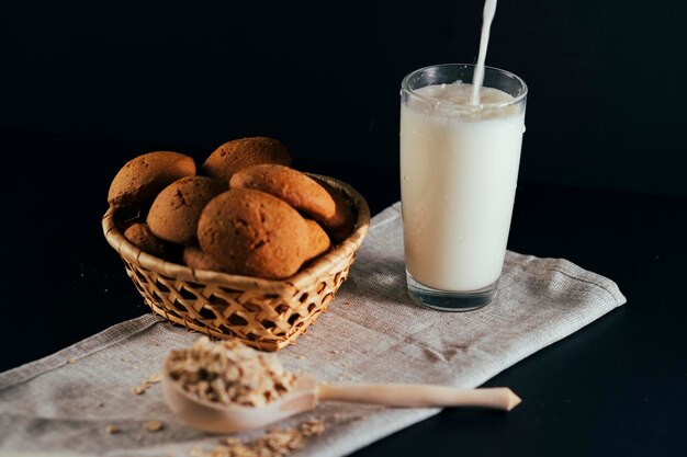 Basket of fresh oatmeal cookies with raw oat flakes in large wooden spoon on napkin on black background Unrecognizable person pouring milk into glass