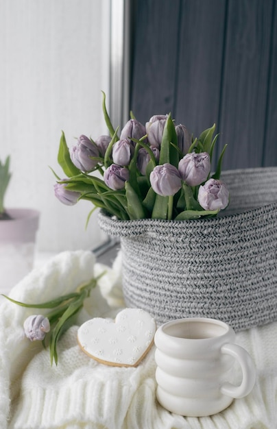 A basket of purple tulips sits on a table with a heart shaped cookie and a cup of coffee.