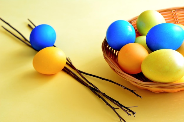 A basket with blue, yellow and orange eggs with twigs on yellow background, close-up