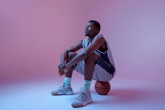 Giocatore di basket seduto sulla palla in studio, sfondo al neon. baller maschio professionista in abbigliamento sportivo che gioca a giochi sportivi, sportivo alto