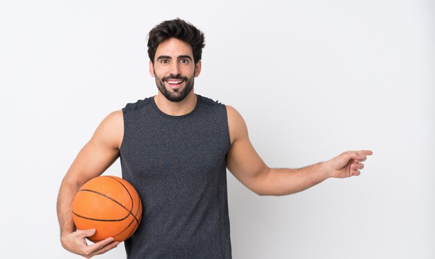 Foto basketbalspeler man met baard over geïsoleerde witte muur verrast en wijzende vinger naar de kant