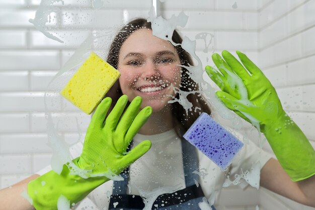 Bathroom cleaning, teenager girl washing shower glass with foam and sponges, closeup of females hand in green gloves and colored sponges