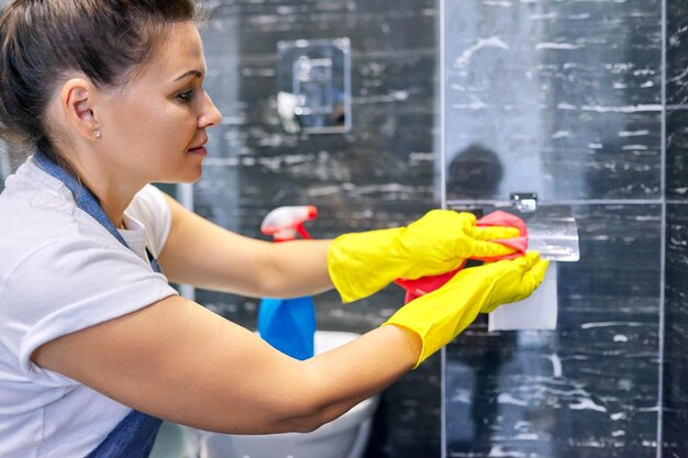 Bathroom cleaning woman in gloves with detergent microfiber rag polishing tiles and chrome accessories