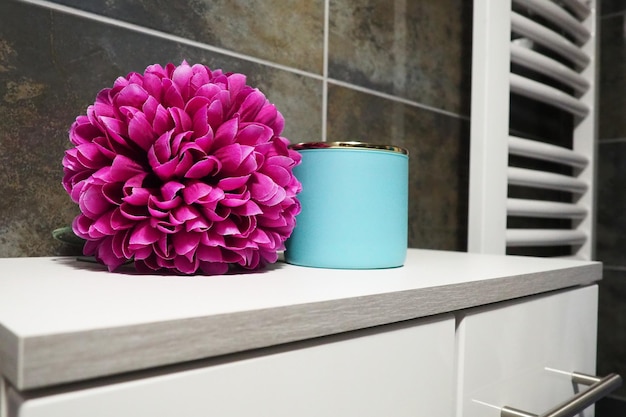 Bathroom decor elements a blue jar of cream and a pink bright\
artificial double flower on a cabinet shelf black wall tiles\
bathroom and toilet interior radiator towel warmer on the wall