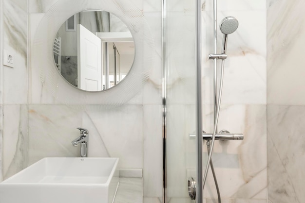 Bathroom tiled in white marble with gray veins circular mirror and shower cabin with glass partition and square sink