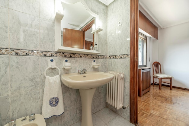 같은 재질의 다리가 있는 베이지색 도자기 싱크대와 흰색 나무 선반이 있는 거울이 있는 욕실