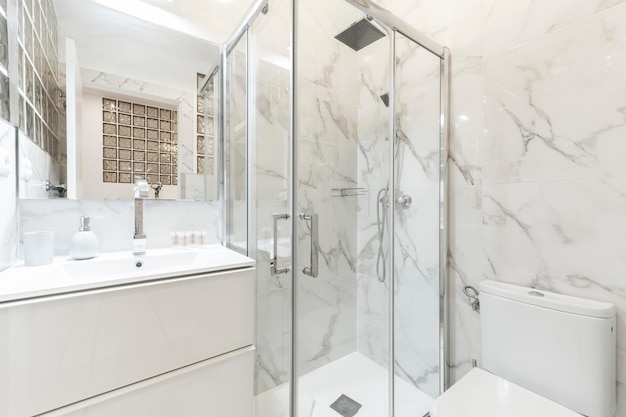 Ванная комната с фарфоровой раковиной, душевым поддоном, прямоугольным безрамным зеркалом и стенами, облицованными белым мрамором с серыми прожилками.