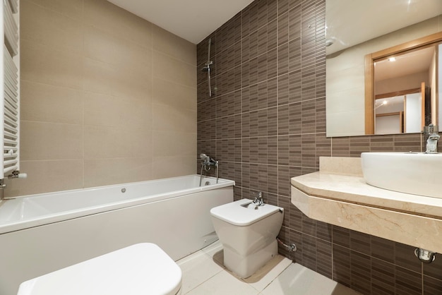 Bathroom with white porcelain sink on cream marble countertop long tub square frameless mirror brown tiles and white heated towel rail