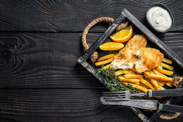 Photo battered fish and chips dish with french fries and tartar sauce in a wooden tray. black wooden background. top view. copy space.