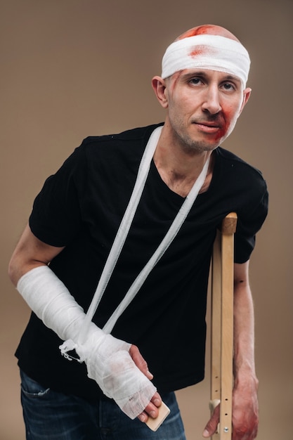 A battered man with a bandaged head and a cast on his arm stands on crutches on a gray background