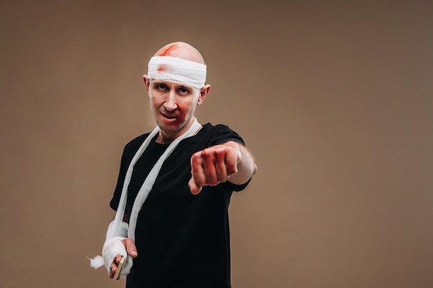 A battered man with a bandaged head and a cast on his arm stands on a gray background.