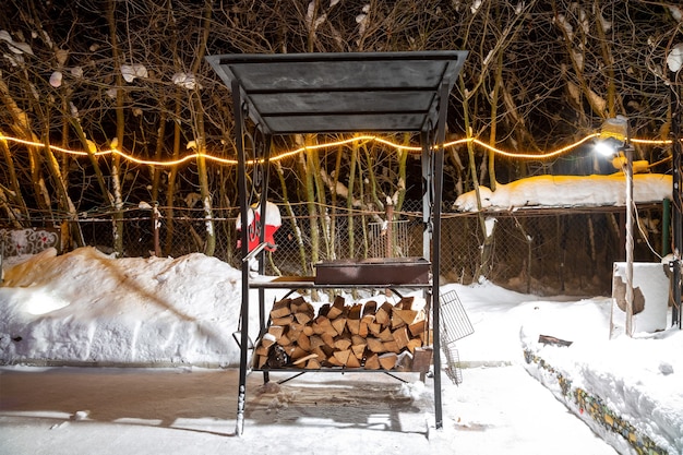 Bbq in de buurt van het huis in de winter. nacht, slingers branden, het sneeuwt. voorbereiding voor het braden van vlees. ze legden geoogst brandhout onder de grill.