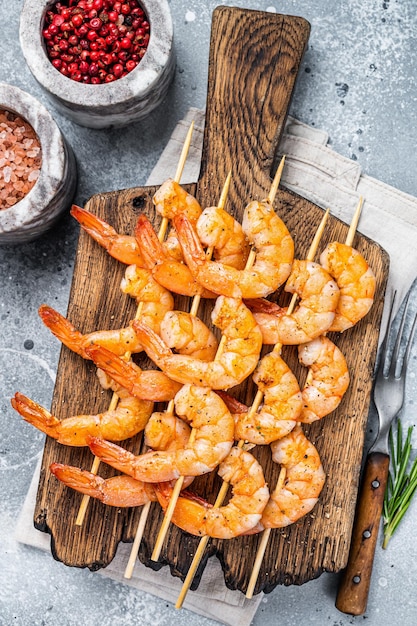 BBQ Roasted shrimps prawns on skewers on a wooden board with herbs. Gray background. Top view.