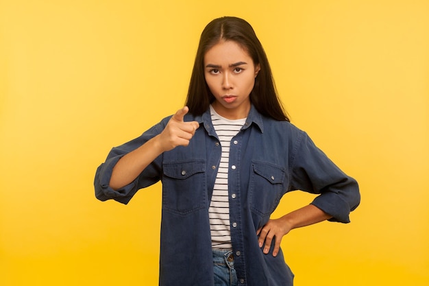 Be careful. Portrait of serious displeased girl in denim shirt pointing finger to camera, warning of mistake, showing disapproval, admonishing gesture. indoor studio shot isolated on yellow background