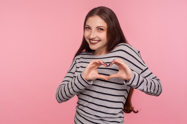 Be my love. Portrait of beautiful happy woman in striped sweatshirt showing heart shape gesture and smiling, showing care symbol, romantic feelings. indoor studio shot isolated on pink background