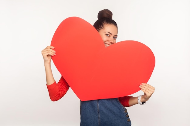 Be my Valentine. Portrait of playful charming happy girl hiding behind big red paper heart and smiling seductively to camera, expressing love, flirting. indoor studio shot isolated on white background