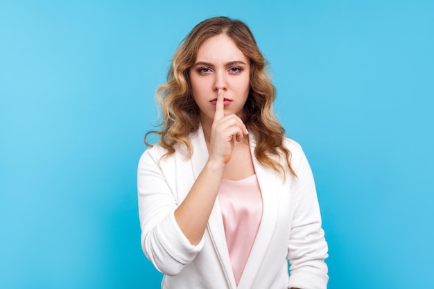 Be quiet, Portrait of displeased angry lady with wavy hair in white jacket making hush sign with finger on lips, demanding silence and secrecy, keep calm. studio shot isolated on blue background